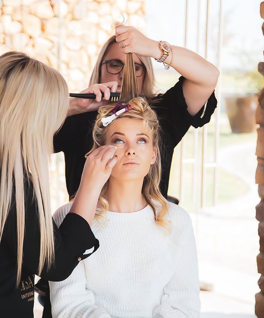 A woman has her hair and makeup done by two stylists. She wears a white sweater, with her eyes closed.