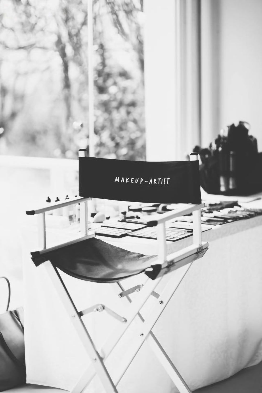 Black and white photo of a makeup artist's chair set up at a table by a window. The chair's back says 