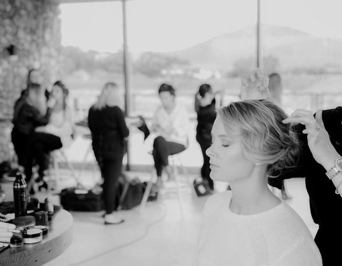 A woman is getting her hair styled with several students in the background, likely a hair and makeup team.