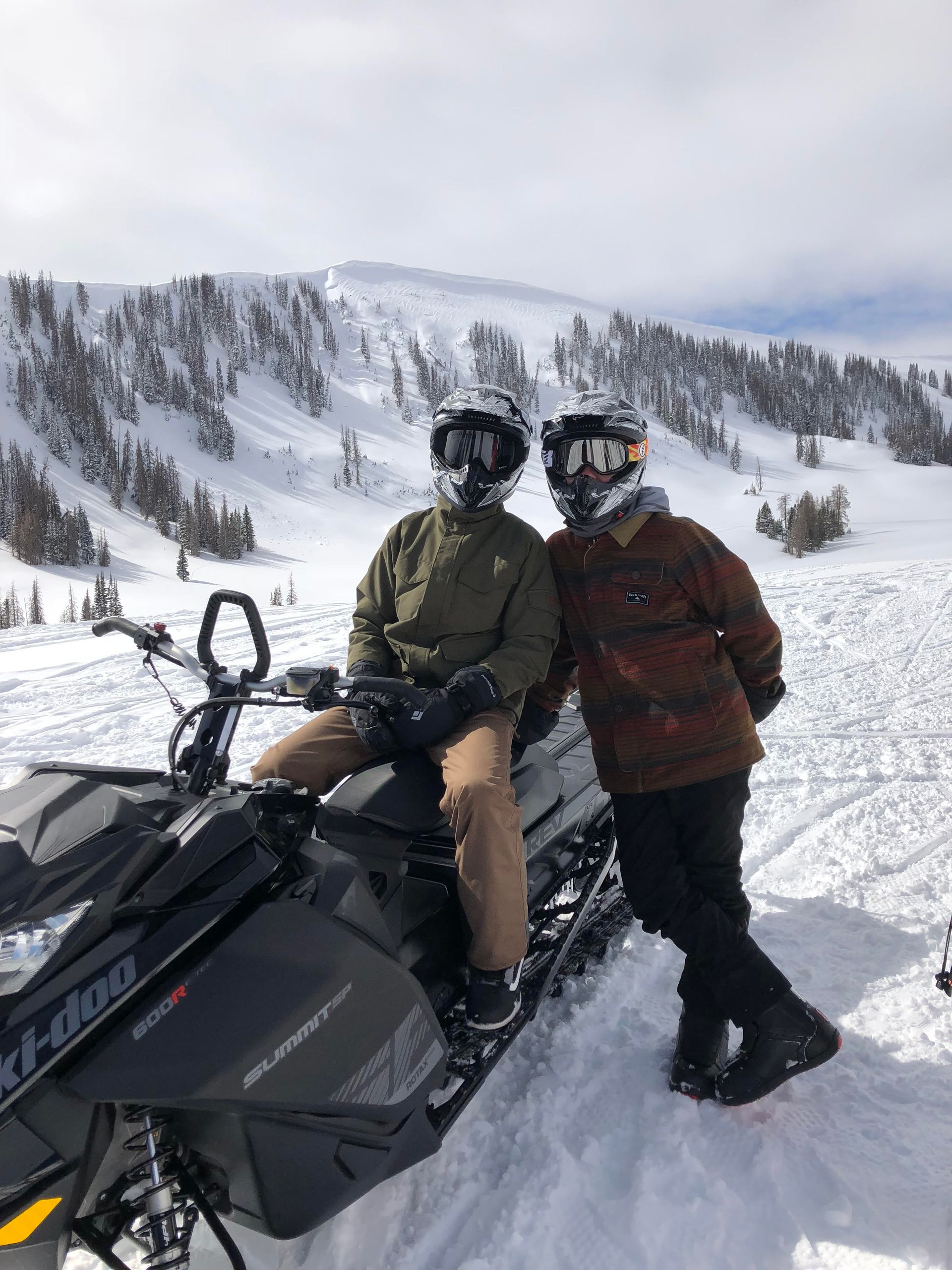 Two people on a snowmobile pose in front of a snowy mountain. One person is seated, the other standing.