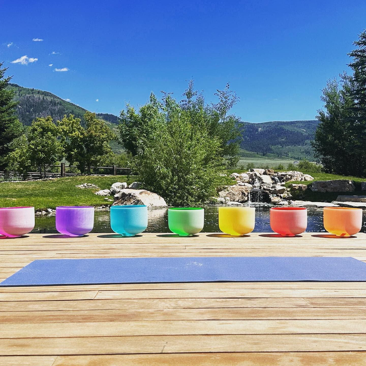 A row of colorful bowls are lined up on a wooden deck next to a yoga mat.