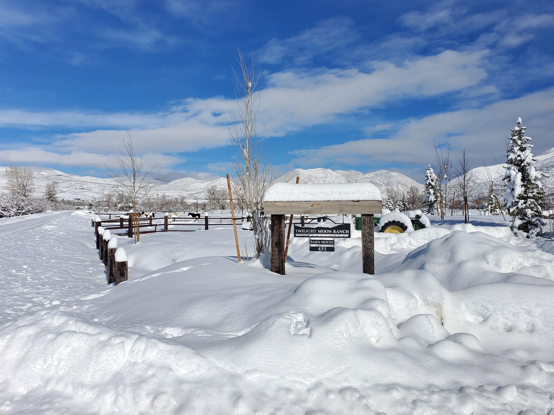 Snow-covered campsite with a sign, picnic tables, and distant snow-capped mountains under a blue sky.
