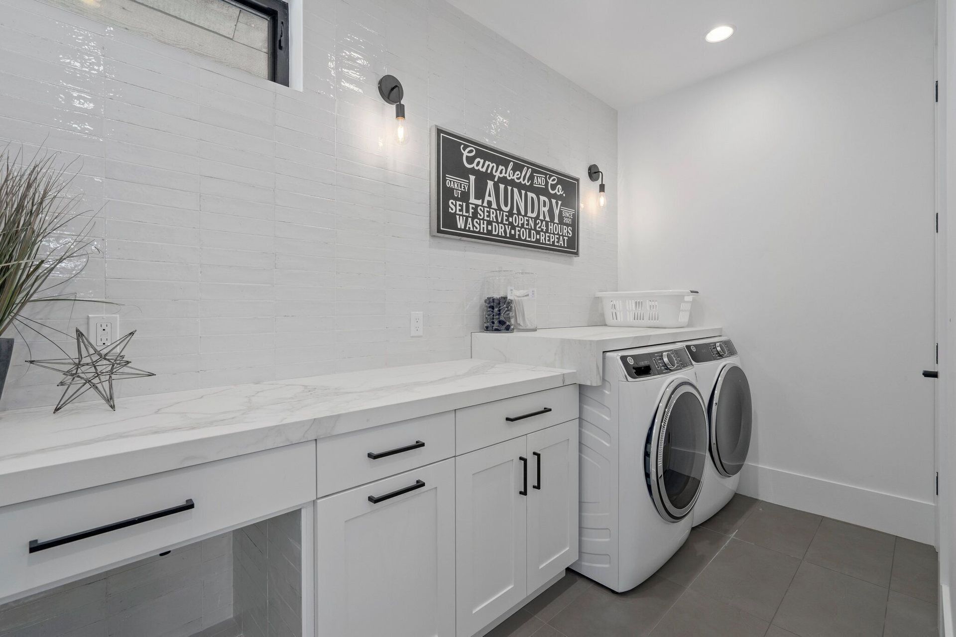 White laundry room with washer/dryer, cabinets, countertop, and decorative sign; grey floor and white walls.