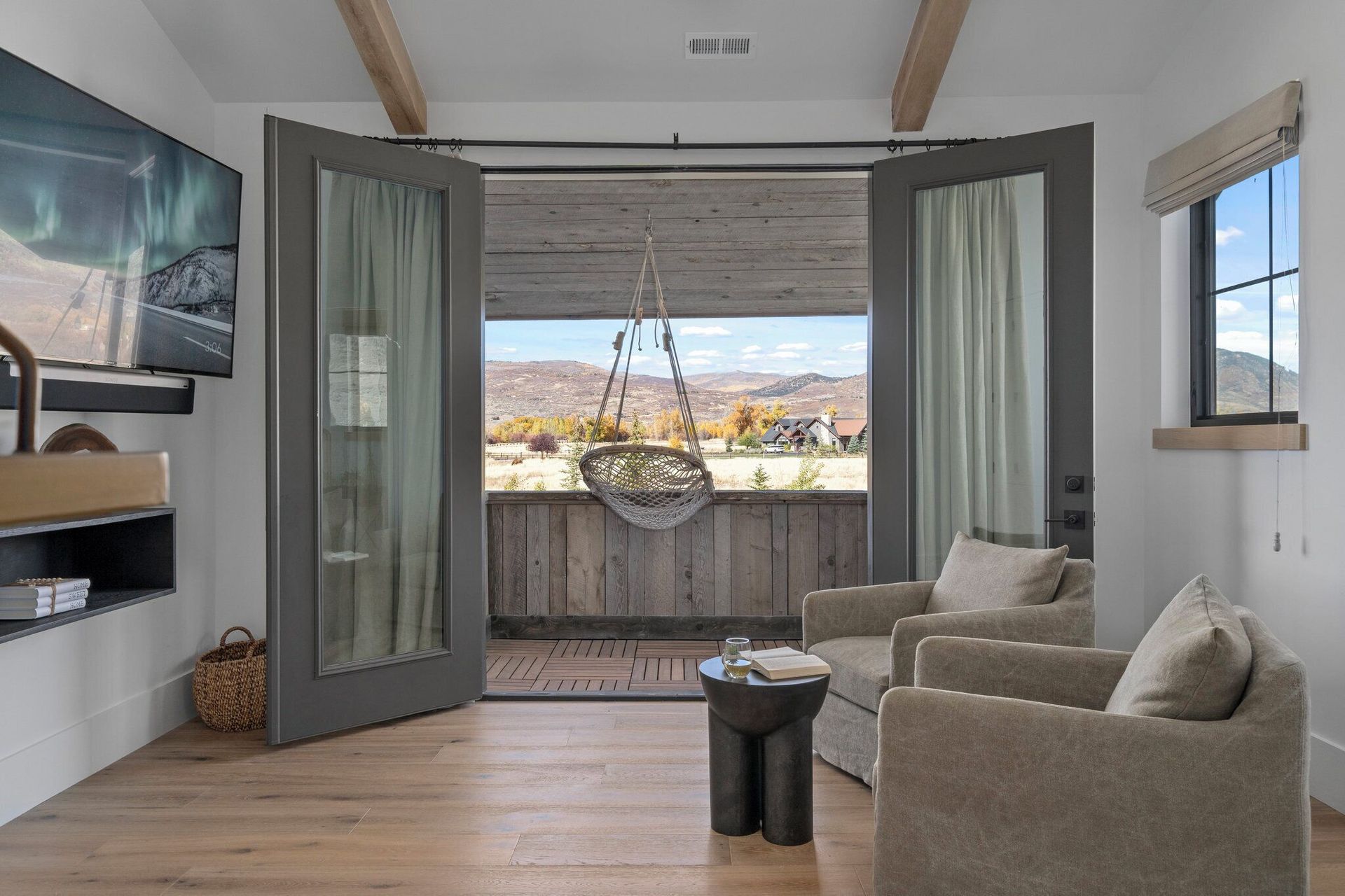 Room with open doors to a balcony with a hanging chair, armchairs, and TV, overlooking a landscape.