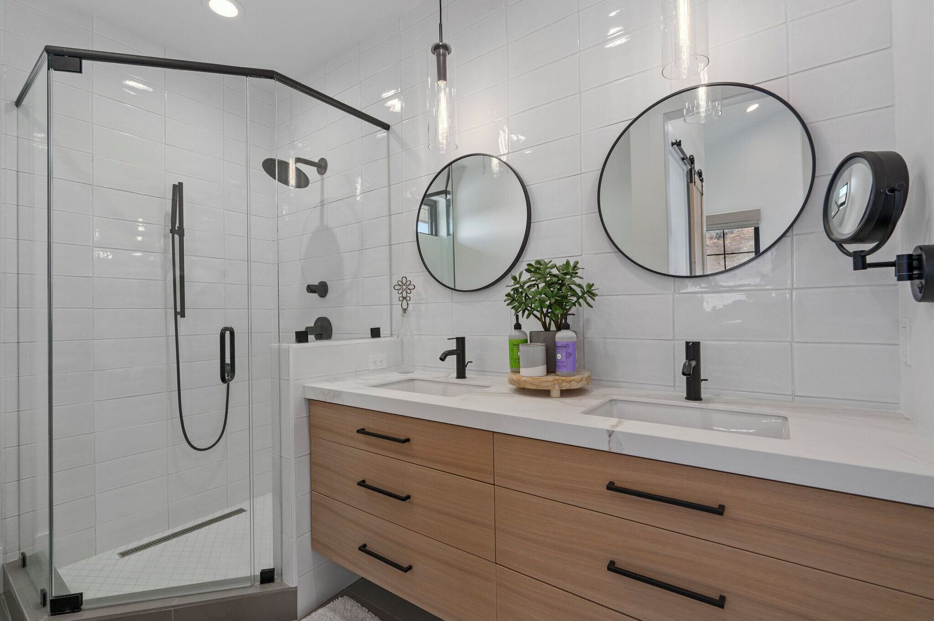 Modern bathroom with wood vanity, glass shower, white tile, black fixtures, and round mirrors.