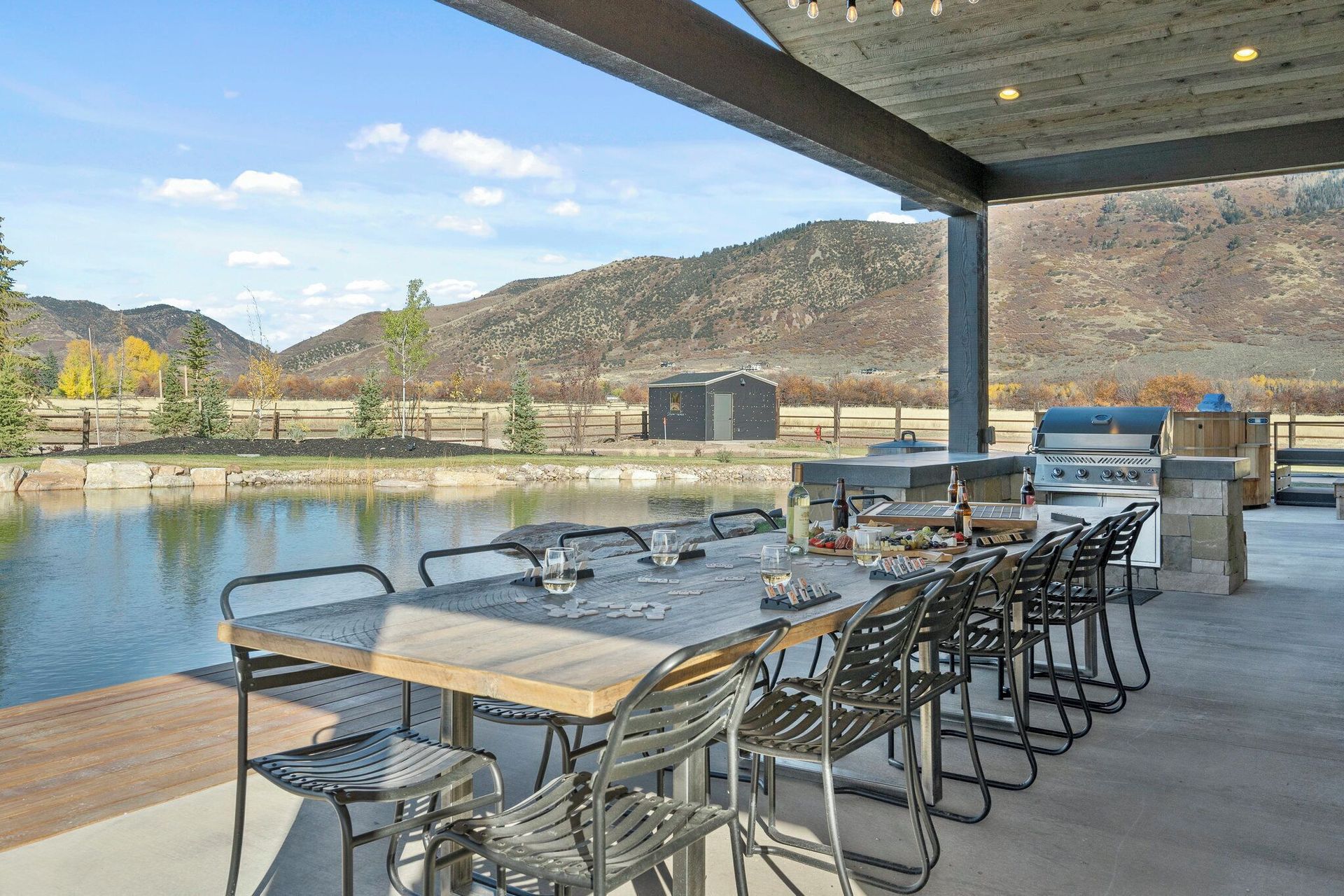 Outdoor patio with a long table, chairs, grills, overlooking a pond and mountains.