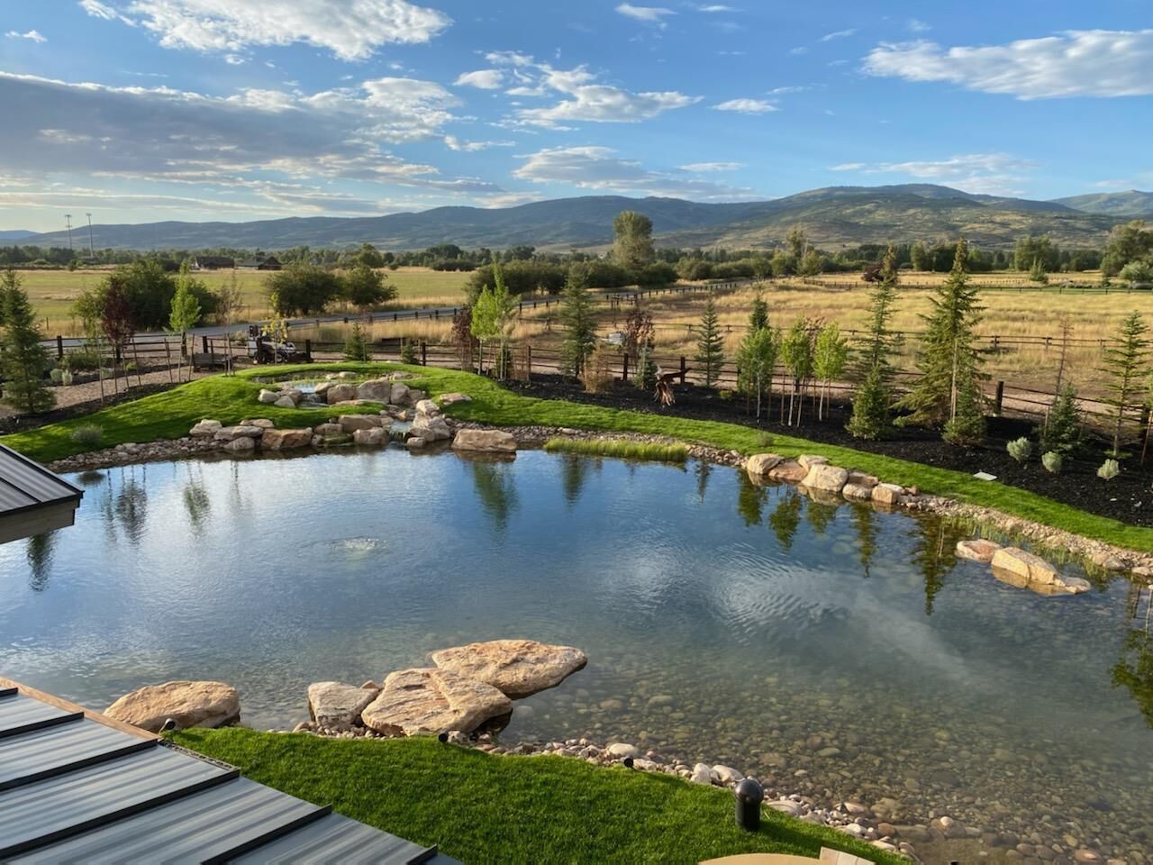 Tranquil pond reflects sky and mountains. Trees and manicured lawn in foreground, fields beyond.