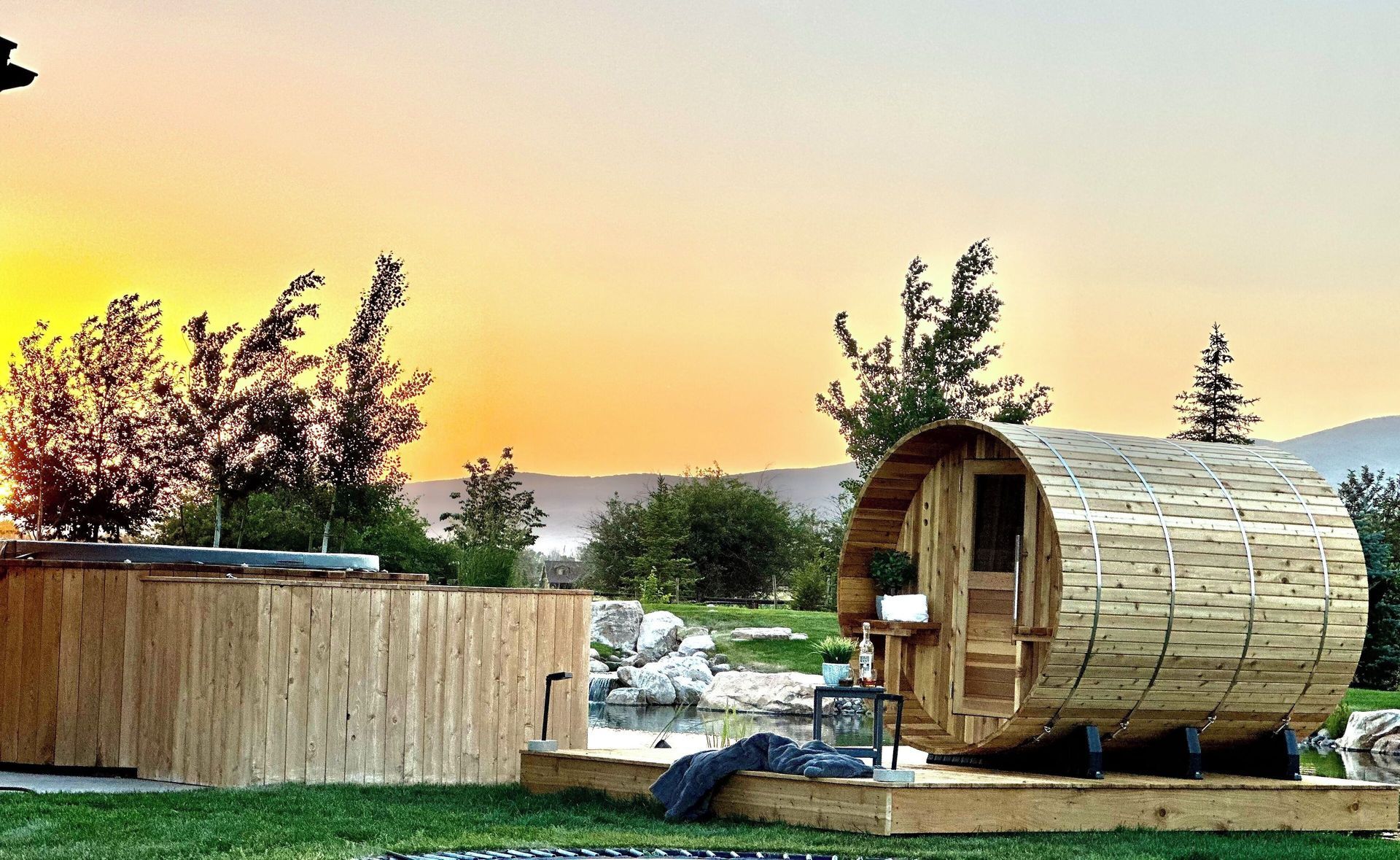 Wooden barrel sauna and hot tub at sunset in a grassy field.