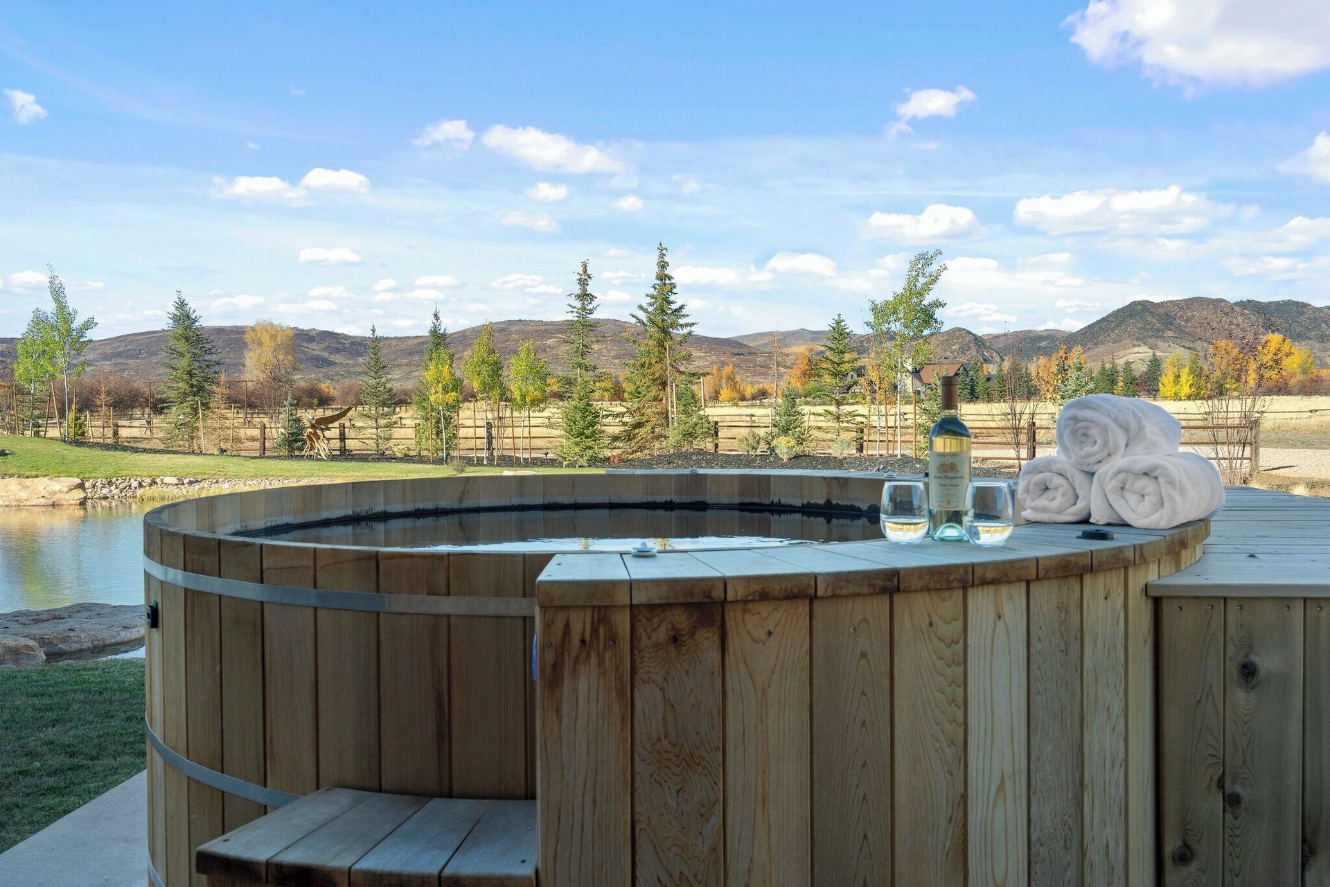 Wooden hot tub outdoors with white wine, towels, and scenic view of trees and hills.