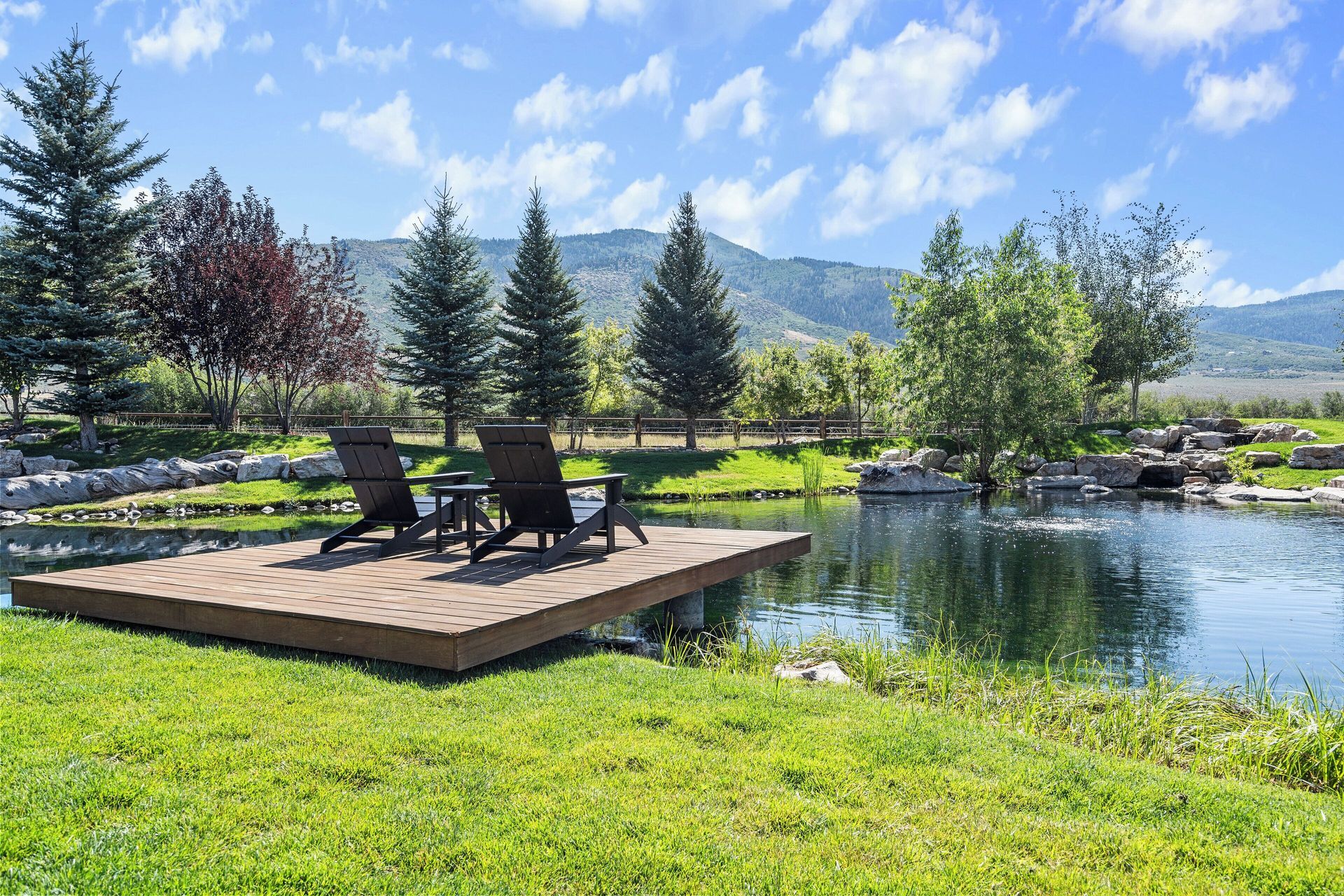Two chairs on a wooden dock over a pond, with mountains in the background.
