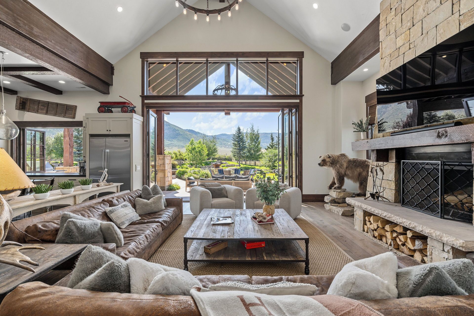 Spacious living room with open doors to a patio. Brown leather sofa, stone fireplace, bear statue, and mountain view.