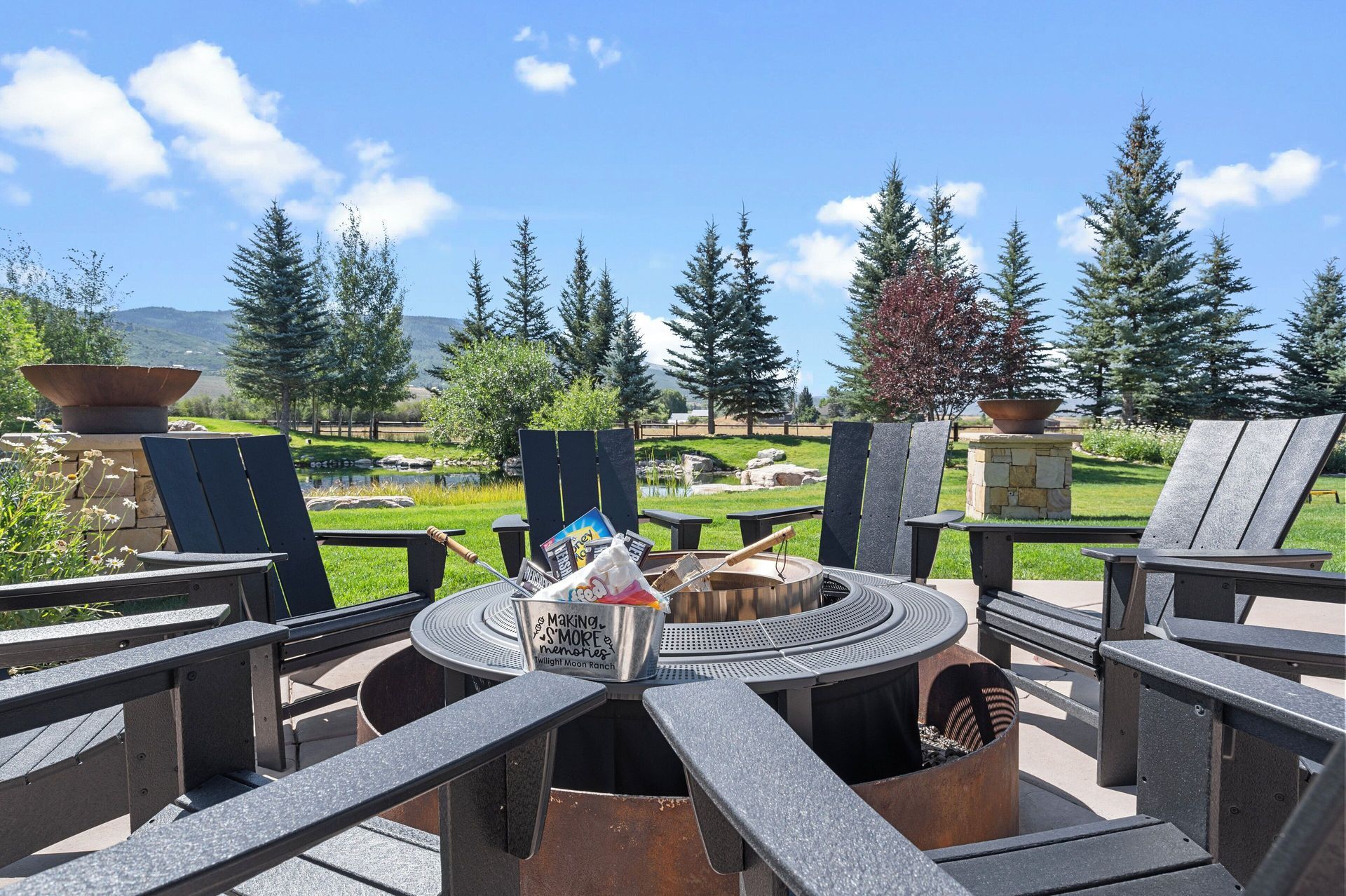 Outdoor fire pit with chairs, surrounded by trees and greenery on a sunny day.