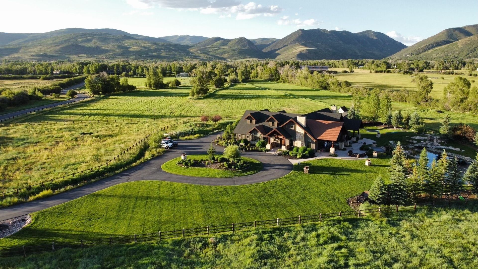 A large house with a circular driveway in a green field, mountains in the background.