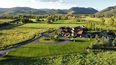 A large house with a circular driveway in a green field, mountains in the background.