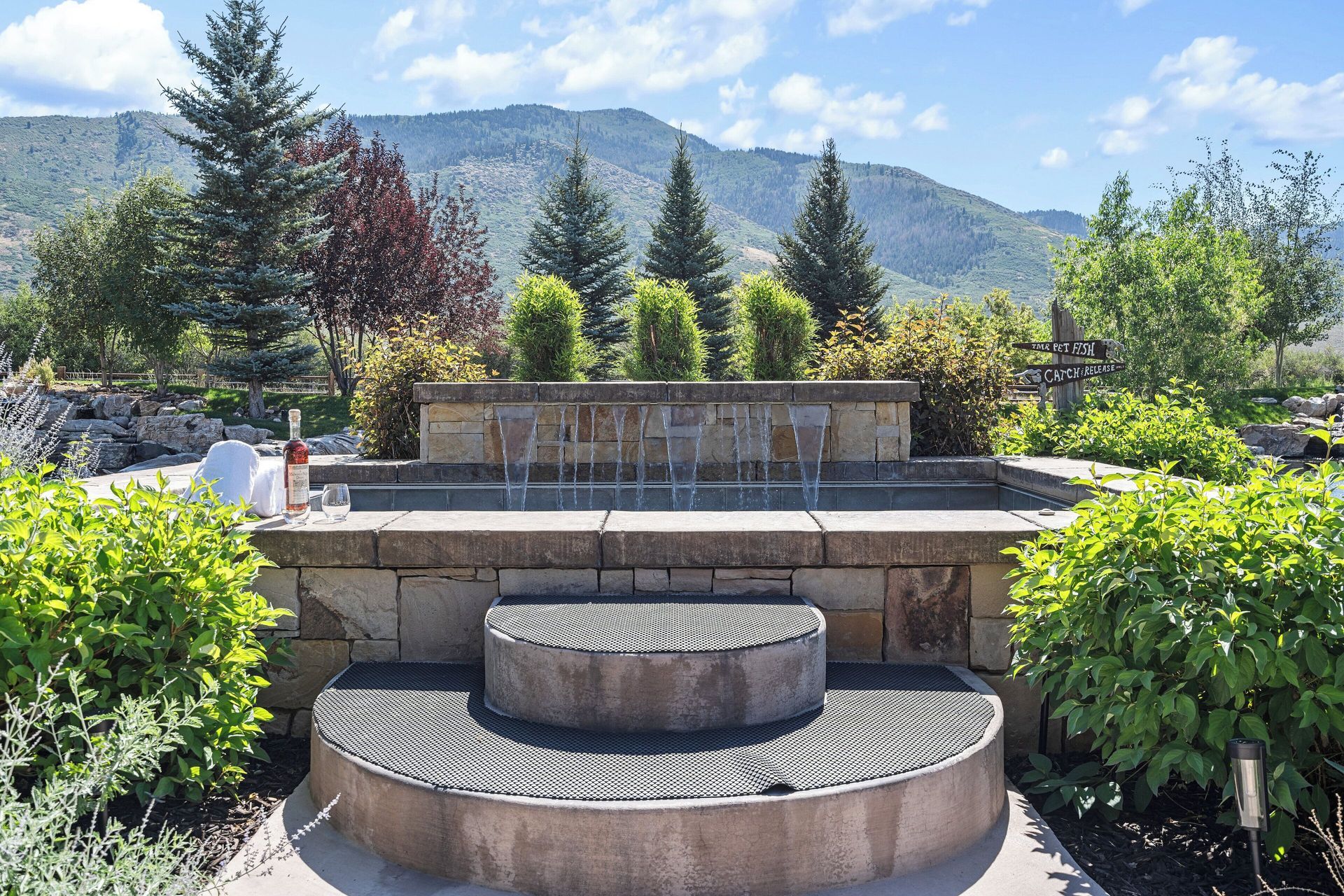 Stone fountain with water cascading down; lush greenery and mountains in the background.