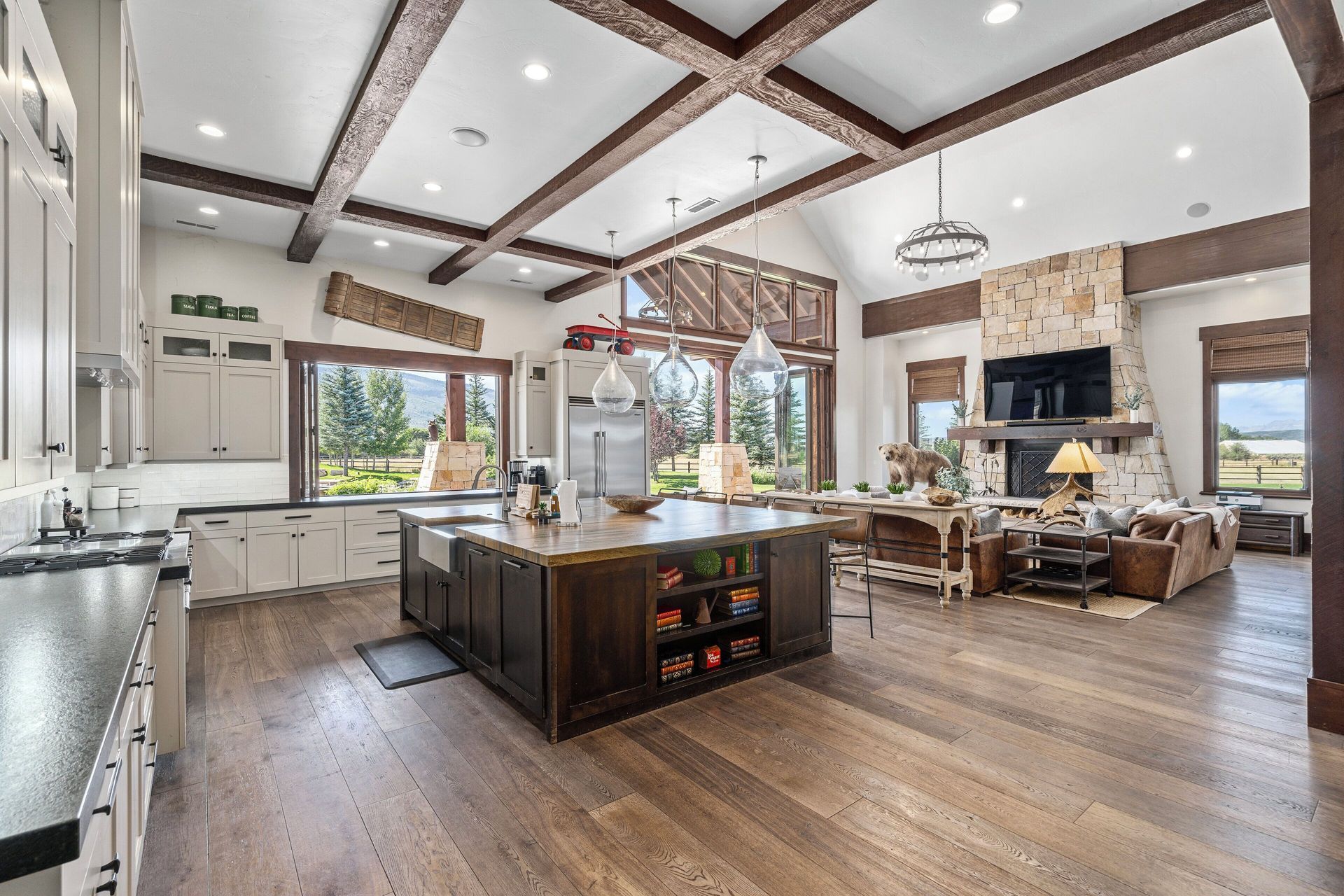 Spacious open-concept kitchen/living room with dark island, wooden beams, large windows, and stone fireplace.