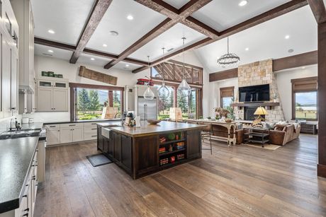 Spacious open-concept kitchen/living room with dark island, wooden beams, large windows, and stone fireplace.