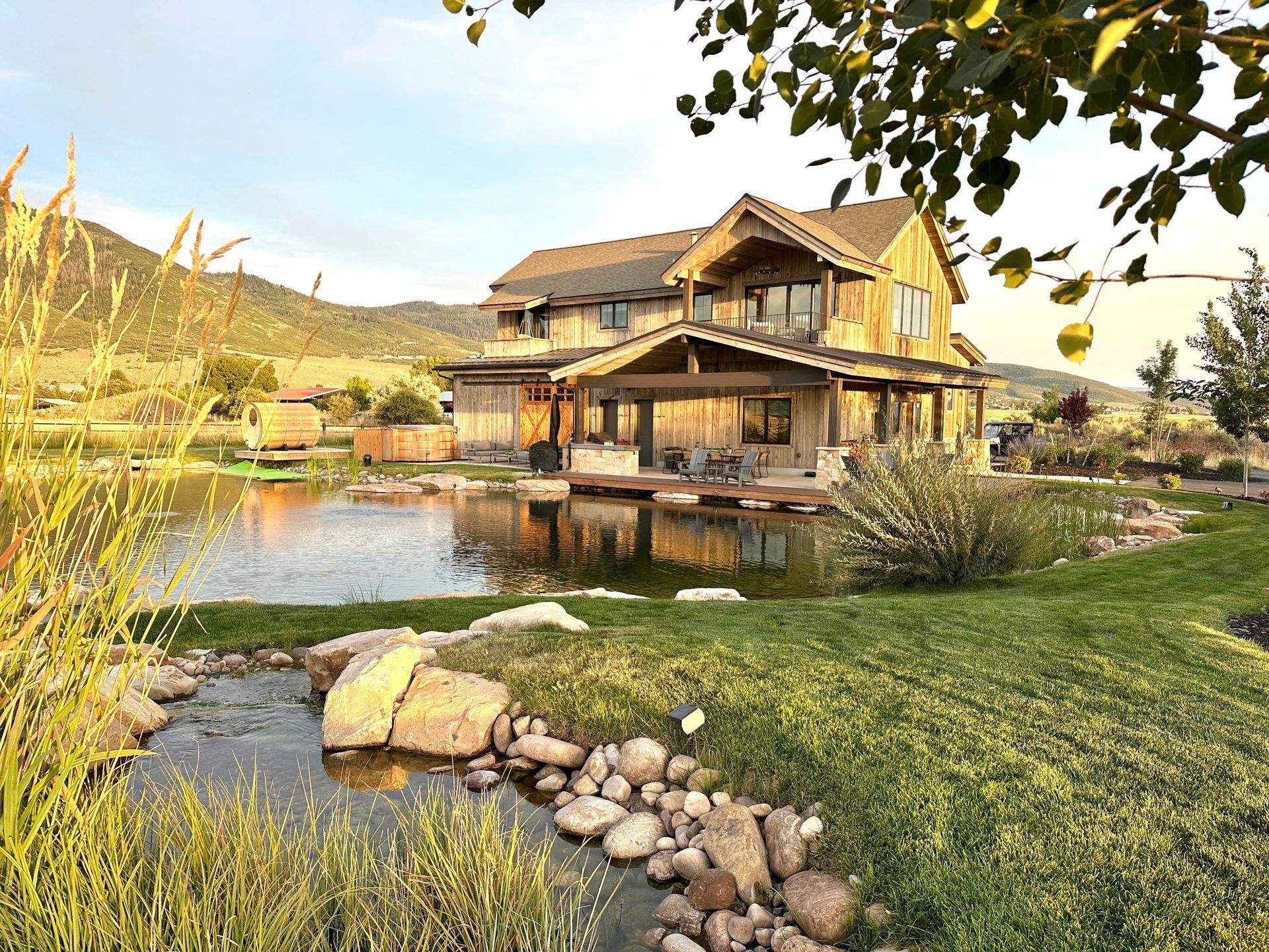 Two-story wooden house with a wrap-around porch sits by a pond. Lush landscaping and a mountain backdrop are visible.