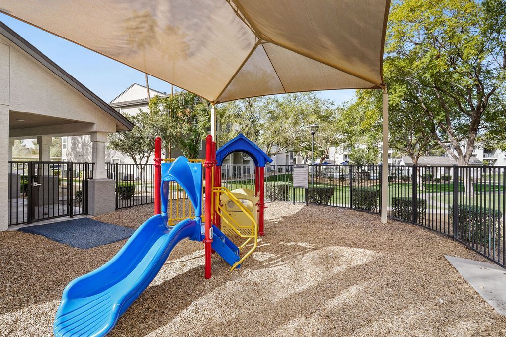 Playground with a blue slide and colorful climbing structure under a shade sail in a gated apartment community.