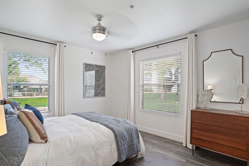 Bright bedroom in a modern apartment with a bed, dresser, mirror, and two windows with blinds.