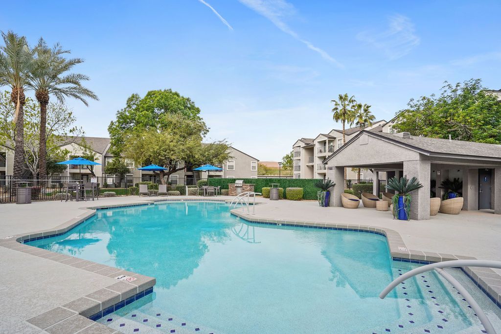Outdoor apartment pool with lounge chairs, blue umbrellas, and palm trees.