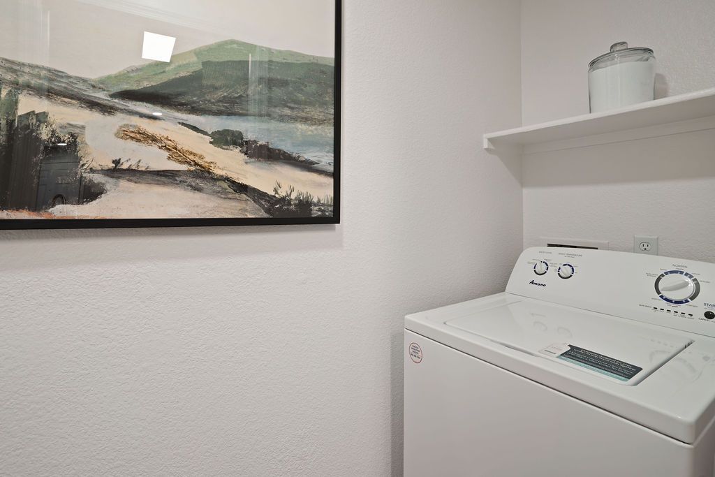 White top-load washing machine in a small laundry closet with a shelf.