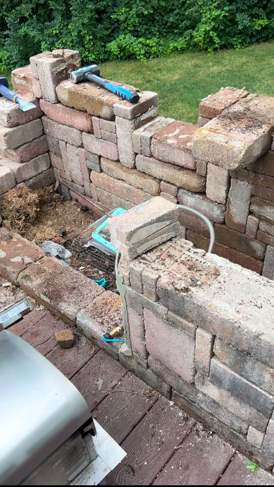 Brick wall construction with tools and debris on a patio, with a grill visible in the foreground.