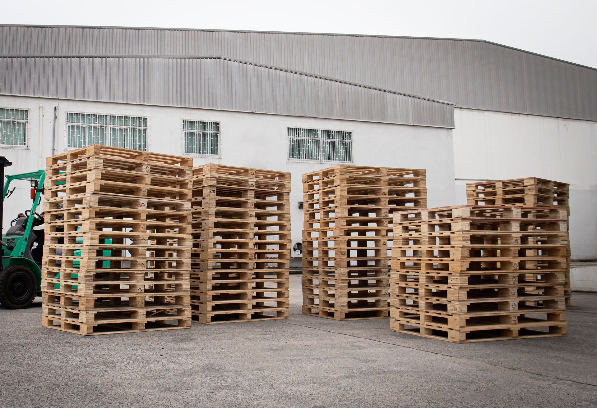 Stacks of wooden pallets in front of a white industrial building. A green forklift is in the left corner.