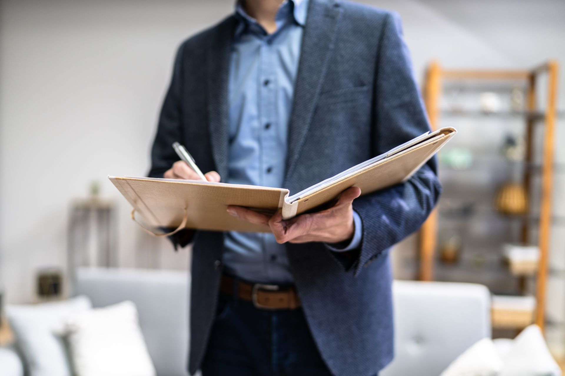 Man in blazer and shirt writing in a notebook indoors, near furniture.