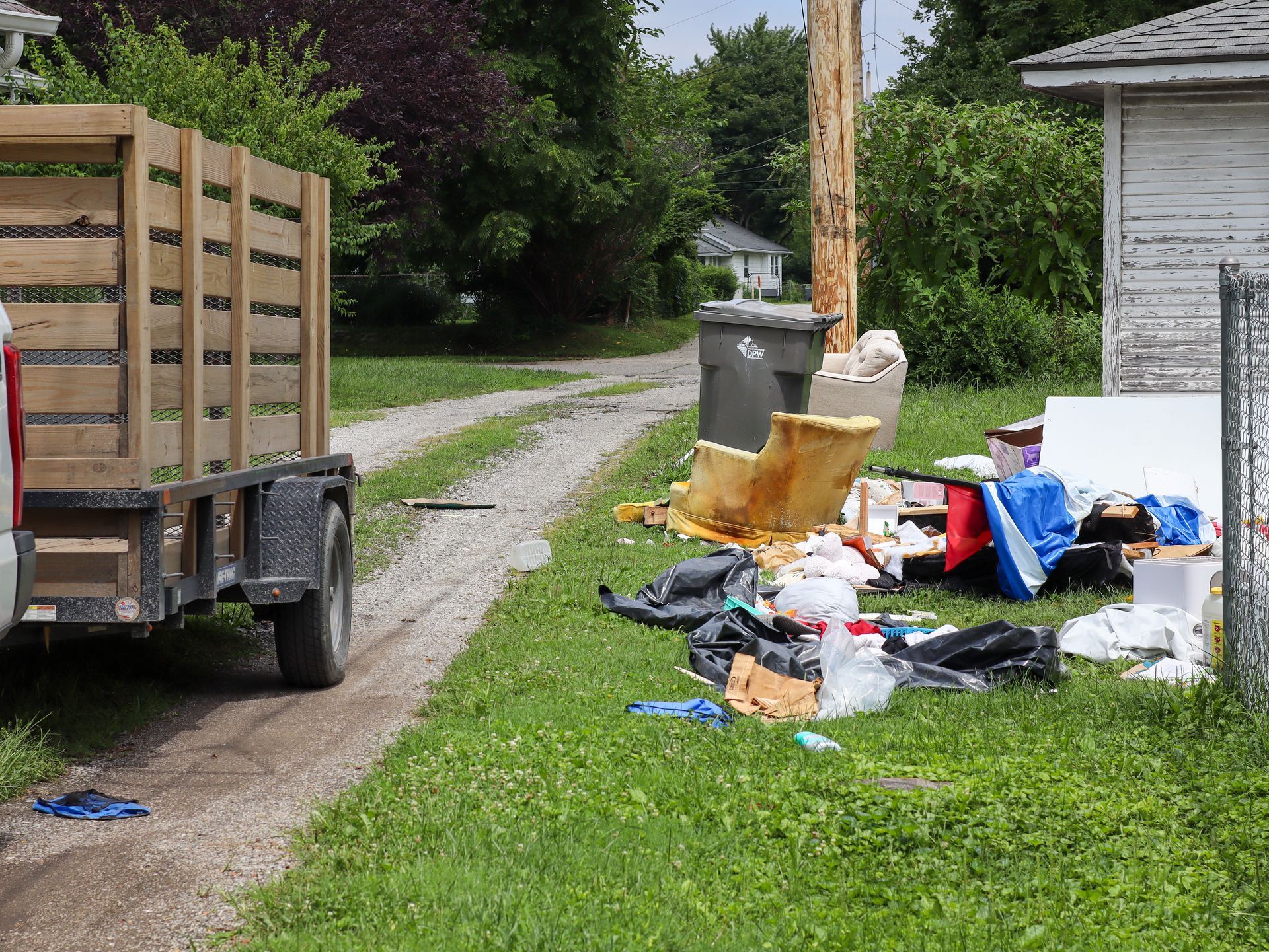Truck with trailer parked near trash pile in a yard; garbage bags, debris, and a damaged trash can.