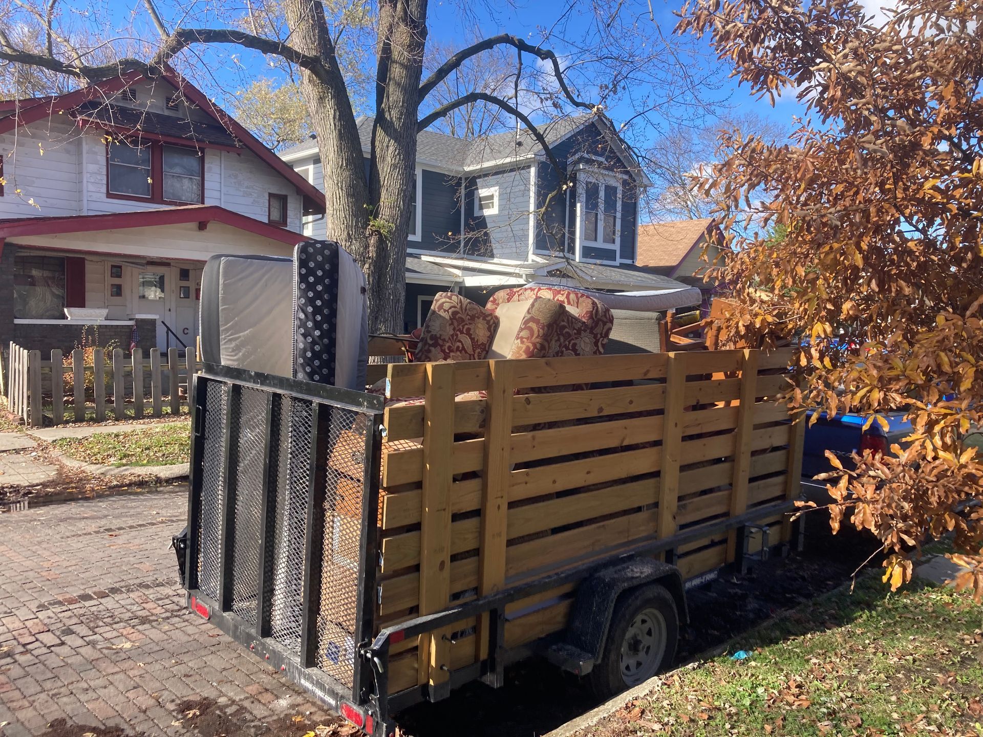 Trailer filled with yard waste and a mattress parked on a residential street with houses and fall foliage.