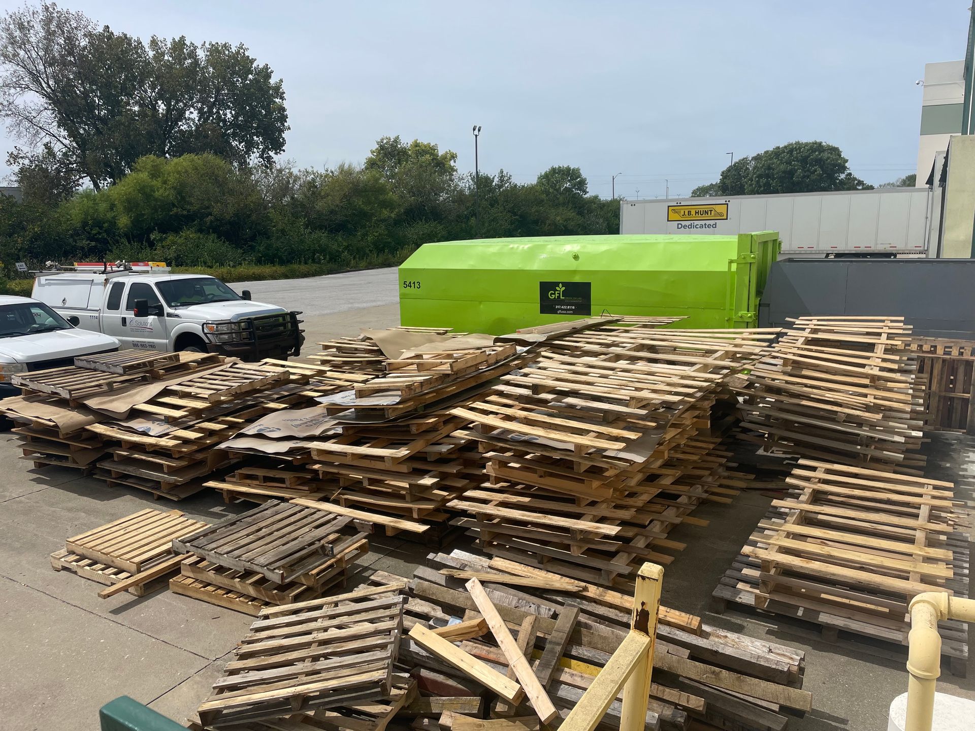 Stacked wooden pallets in an outdoor lot beside a bright green dumpster and parked trucks.