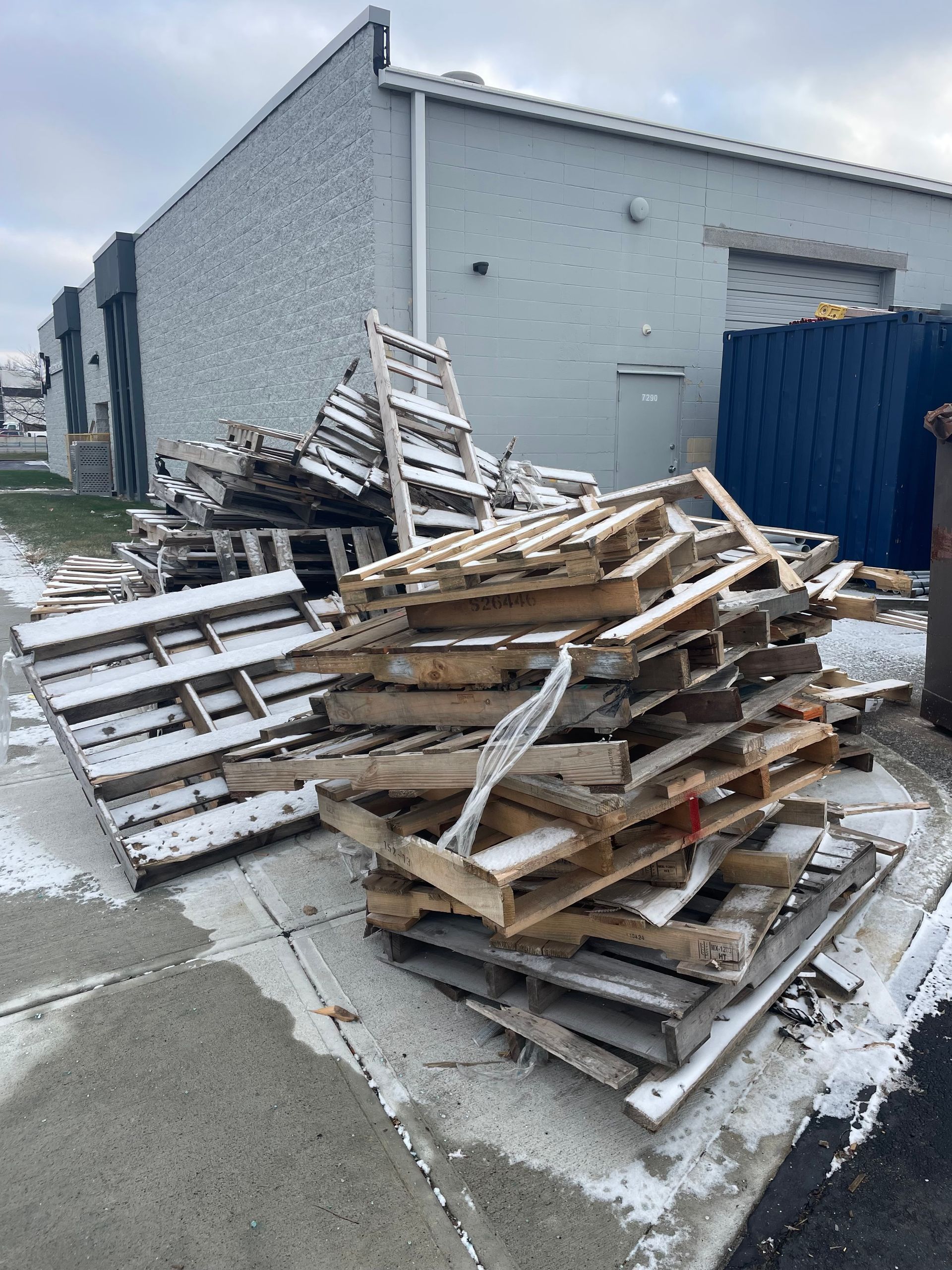 Pile of wooden pallets covered in snow, next to a building and dumpster on a sidewalk.