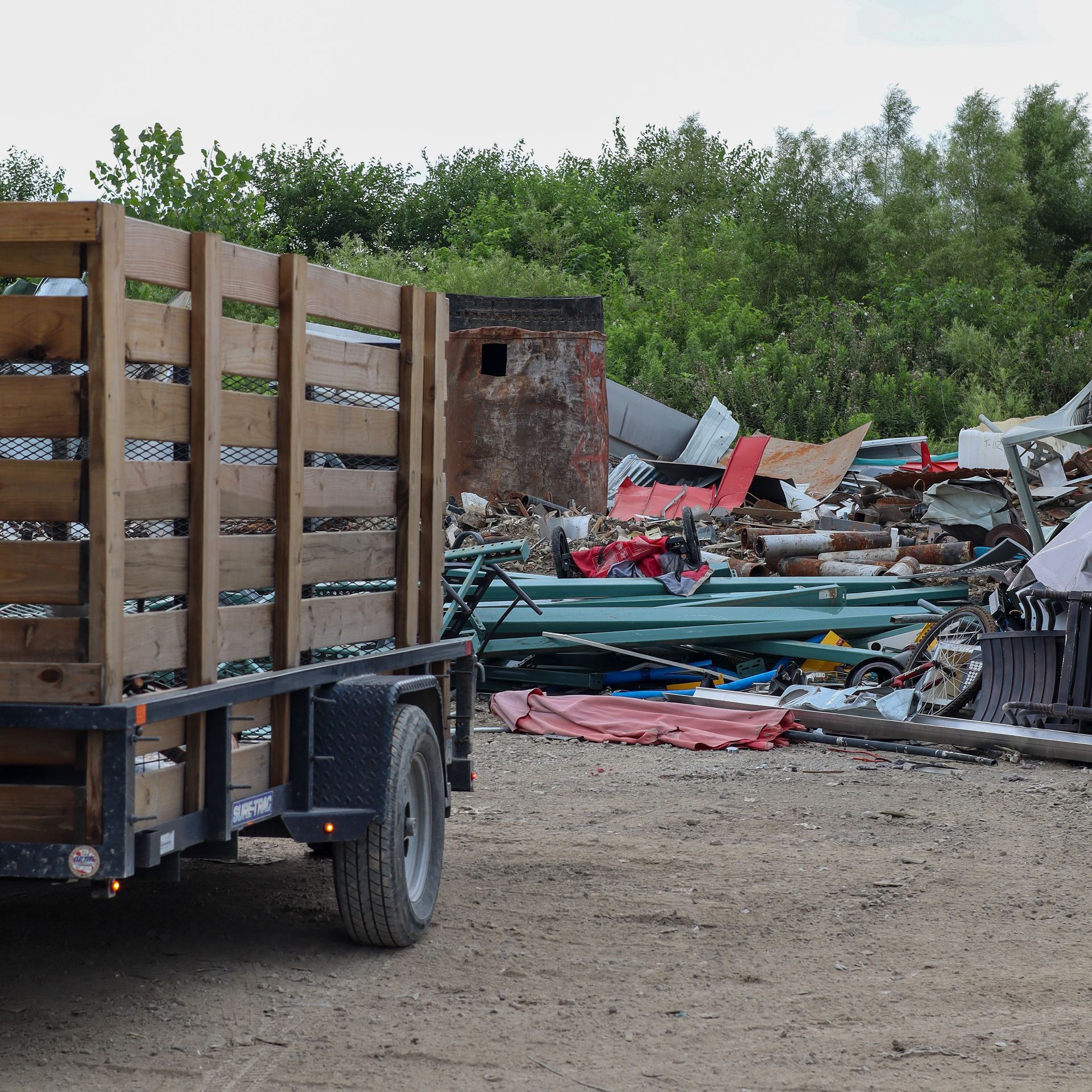 Trailer loaded with wood crates parked in front of a pile of scrap metal and debris in a wooded area.