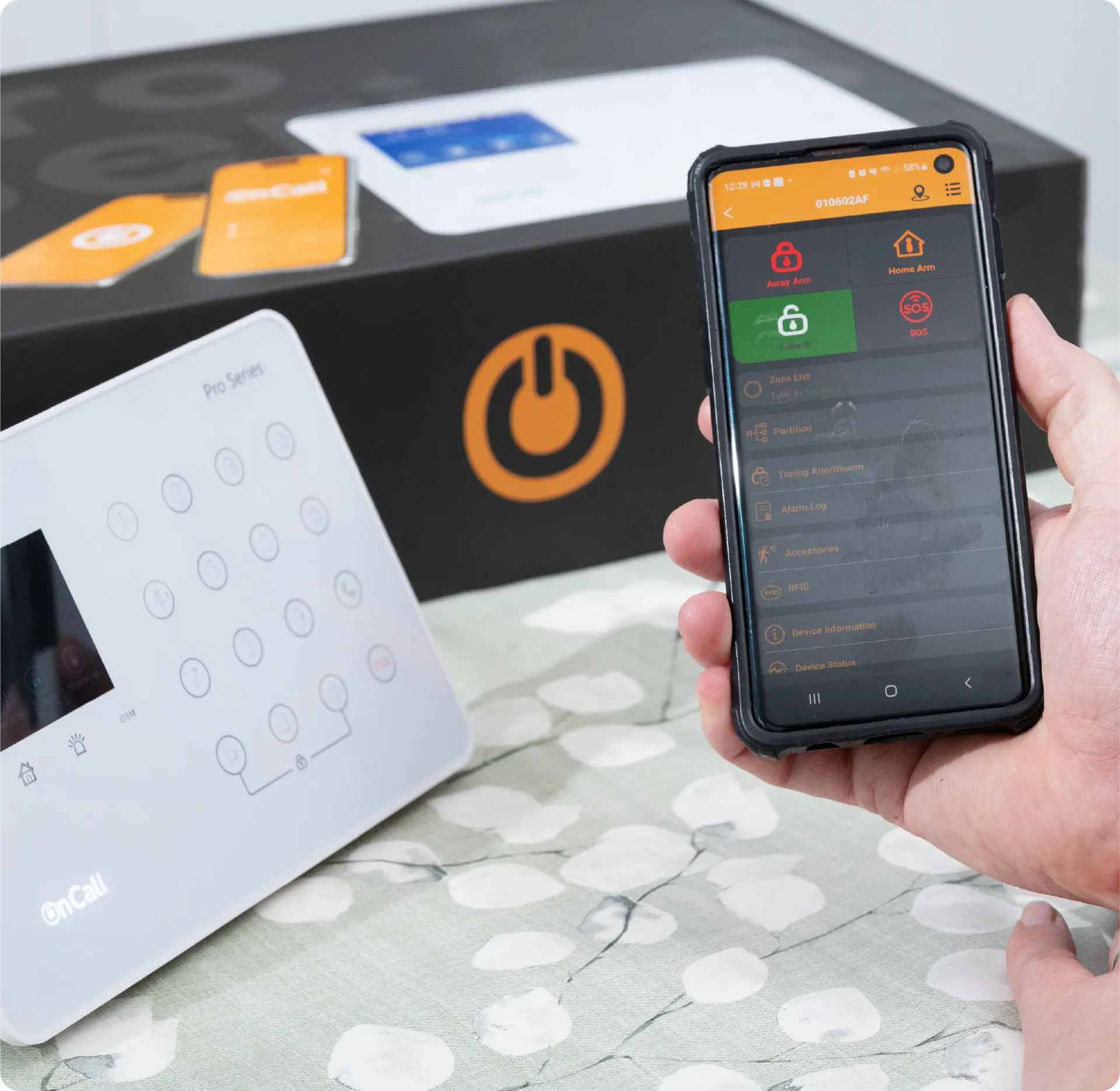 A person uses a smartphone to control a security system. A keypad and product box are also visible.