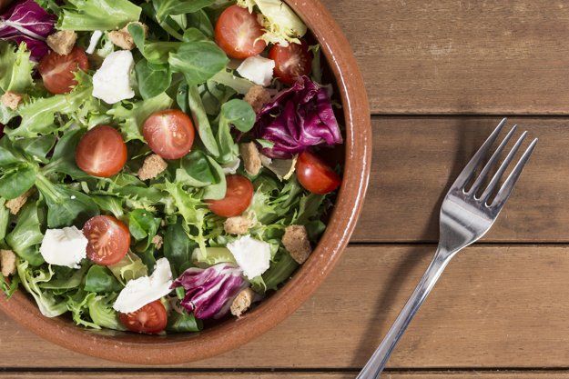 Bowl of salad featuring mixed greens, sliced cherry tomatoes, purple cabbage, croutons and meat. Bowl is on a wooden table with a fork to the right.