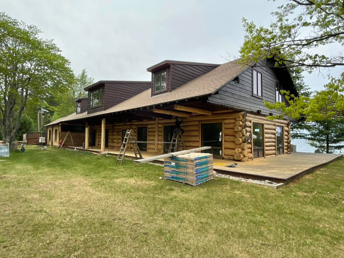 Log cabin under construction with a deck, surrounded by green grass and trees.