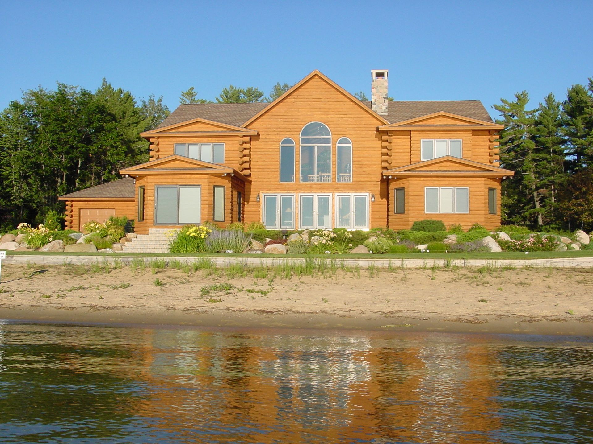 Large log home on sandy shoreline reflecting in water.