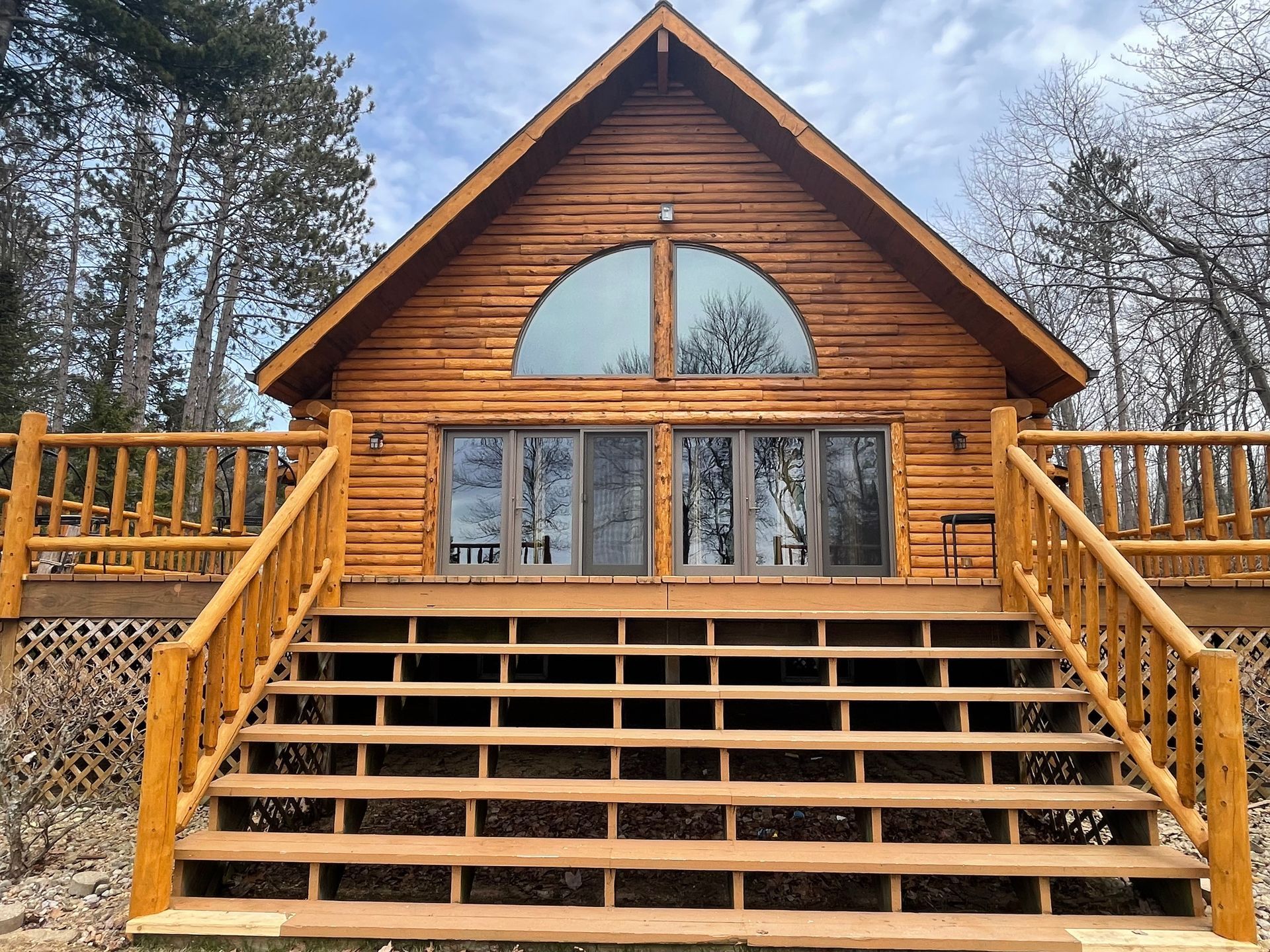 Wooden cabin with large front deck and staircase. Tall arched window above sliding glass doors.