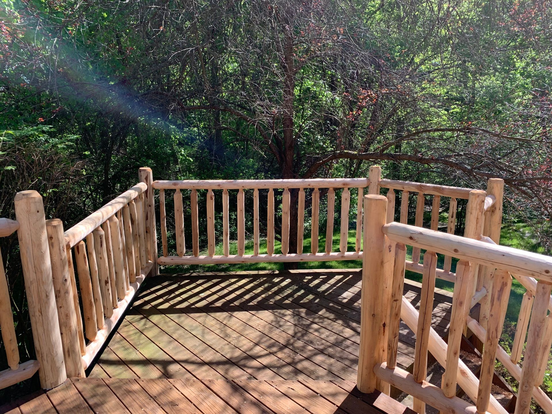 Wooden deck with log railing overlooking a green forest. Sunlight streams through trees.
