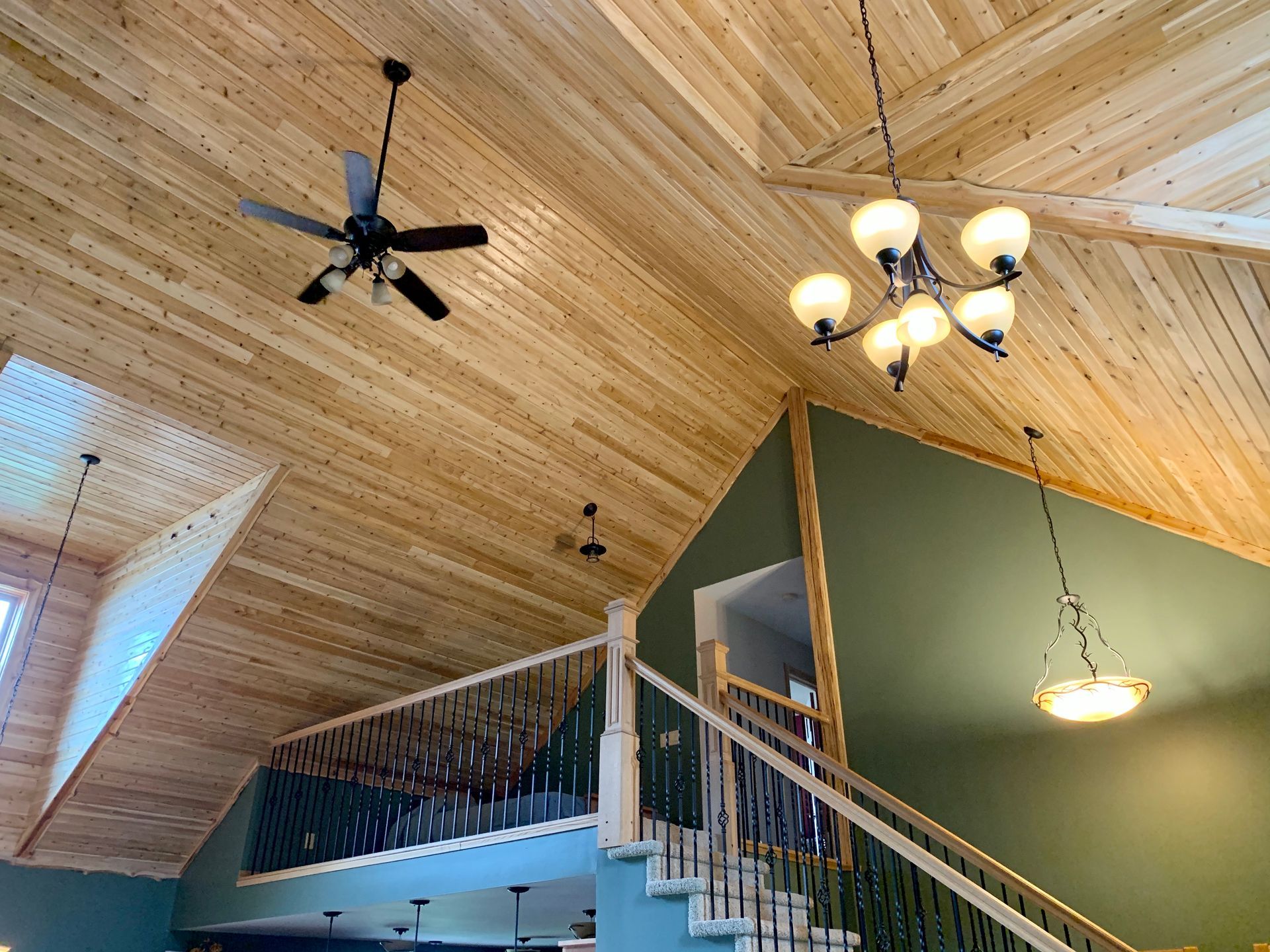 Wooden vaulted ceiling with a fan, chandelier, and staircase against a green wall.