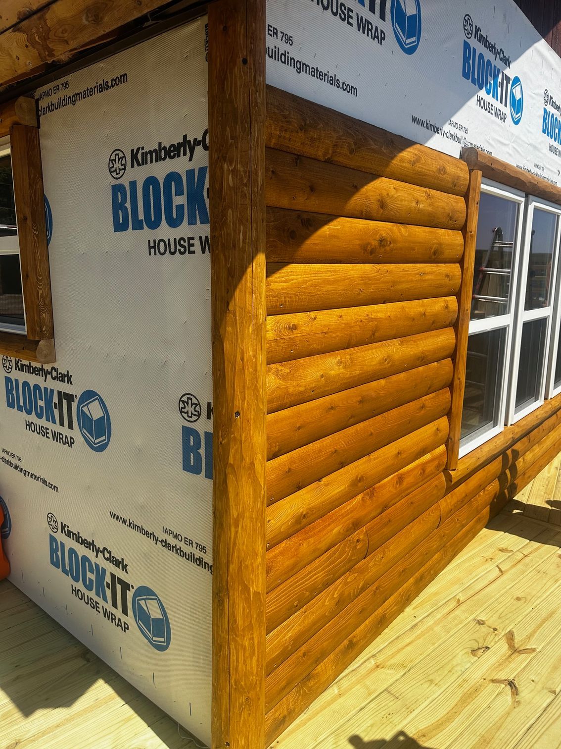 Corner of a cabin with wooden siding, windows, and white house wrap.