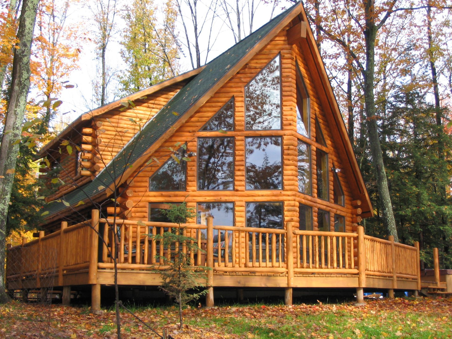A-frame log cabin with large windows and a wraparound deck in a wooded area during fall.