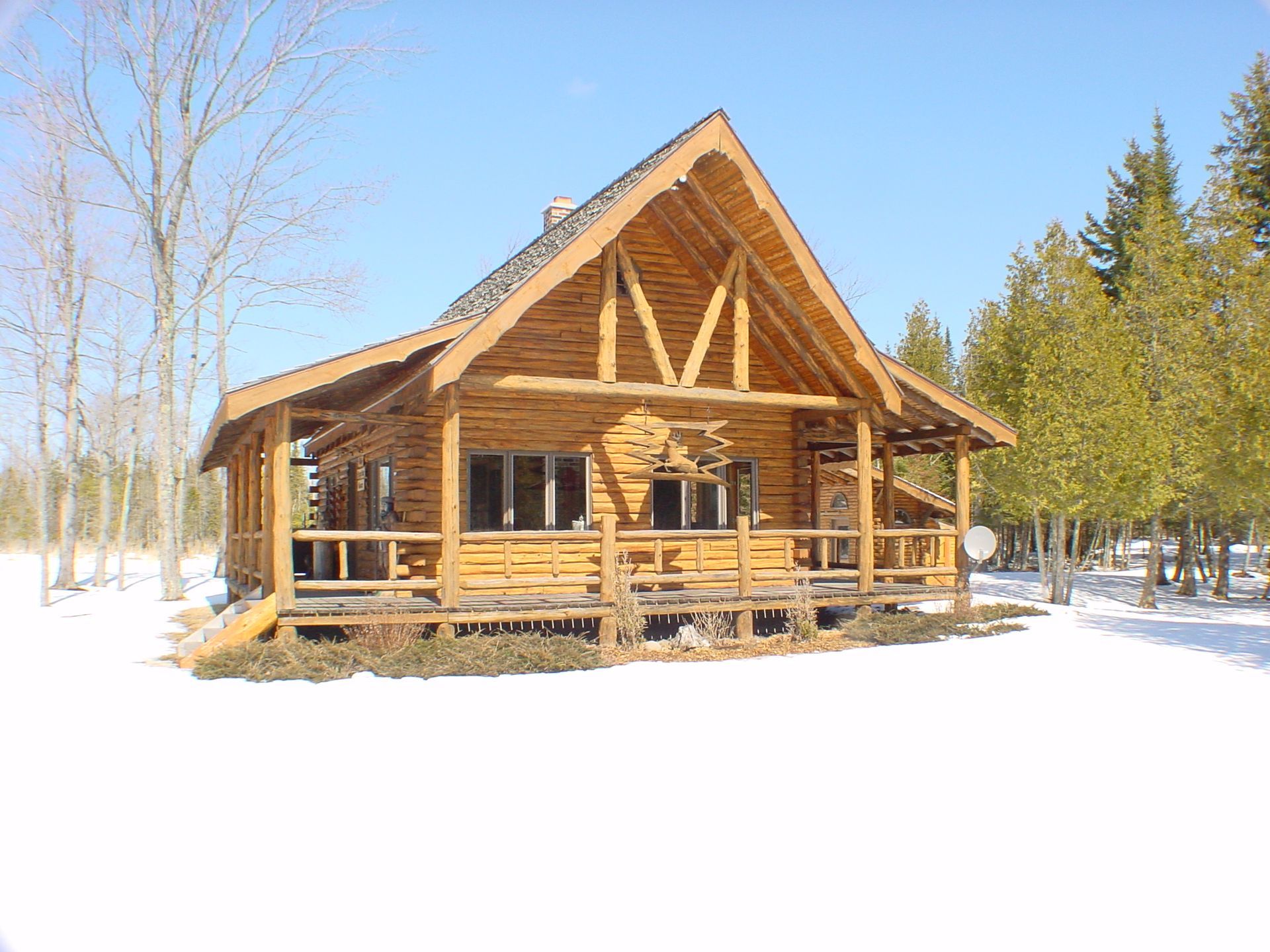 Log cabin on a snowy landscape; wood structure with porch, tall trees, and clear blue sky.