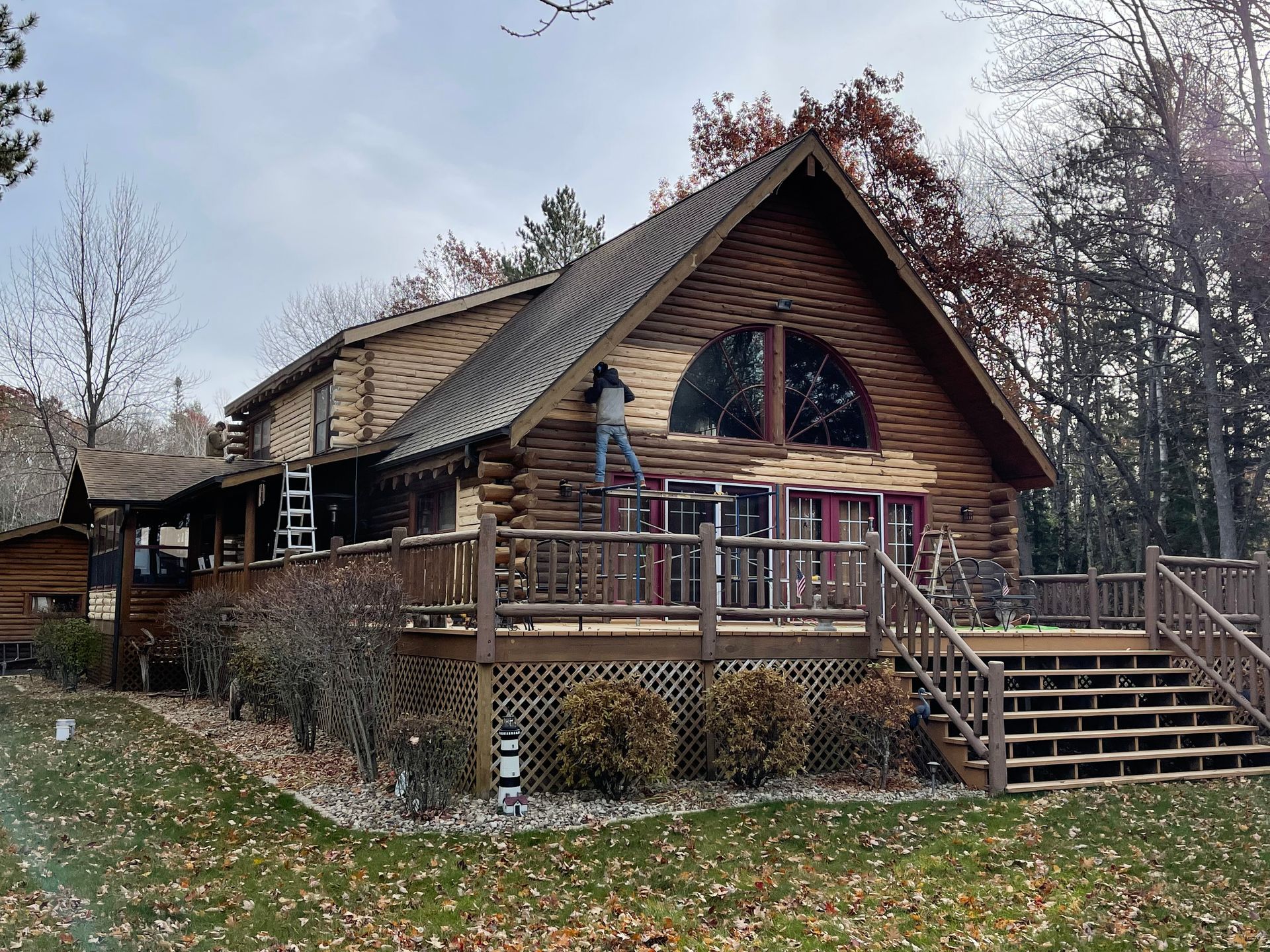 Log cabin with person on ladder, working on the exterior. Wooden deck, lawn, and trees. Overcast day.