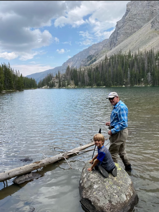 A man and a boy are fishing in a lake while sitting on a rock.