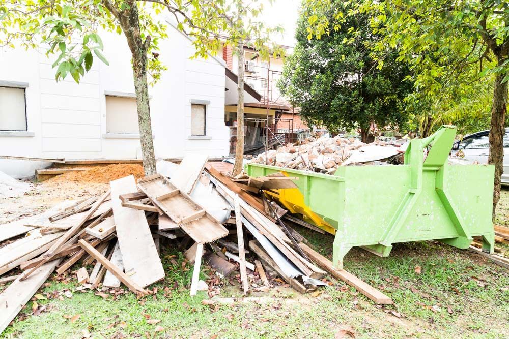 A Green Dumpster Is Sitting Next To A Pile Of Wood In Front Of A Building — Ballina Demolitions In Alstonville, NSW