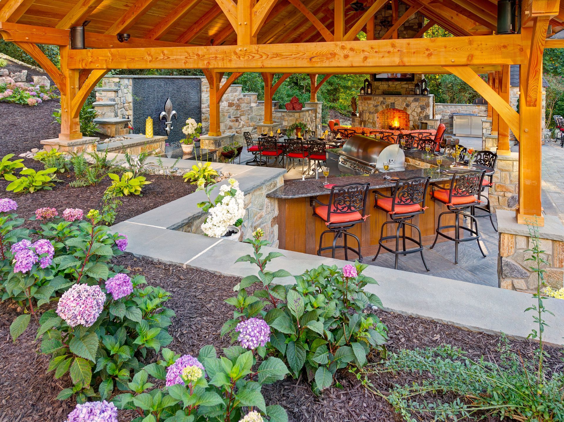 A wooden pavilion with a bar and a fireplace in the backyard surrounded by flowers.
