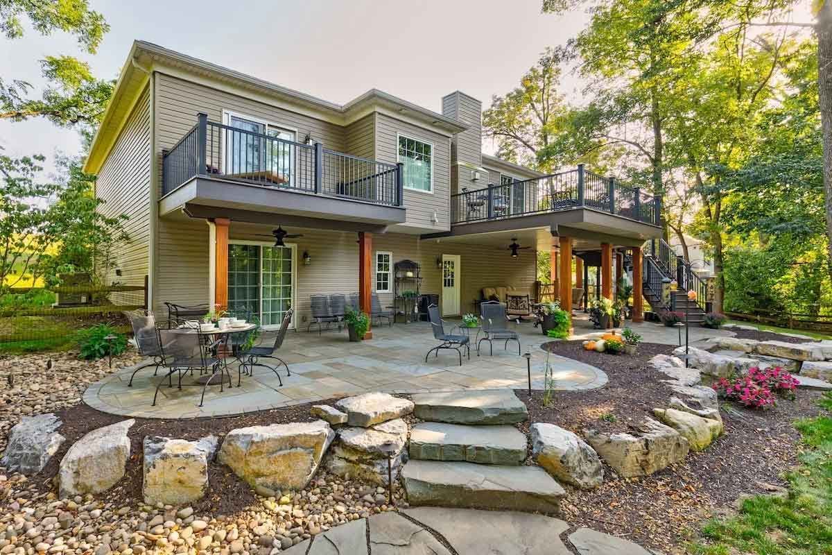 Back patio with two-story house. Stone patio and rock landscaping. Second story balcony, dining set. Sunny, outdoor.