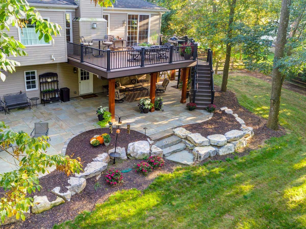 A backyard with a raised deck, stone patio, and landscaping.